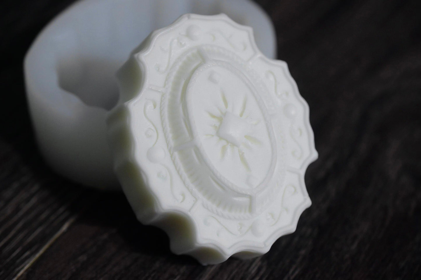 A white, oval soap bar with elegant and filigree design sits next to a white mold.