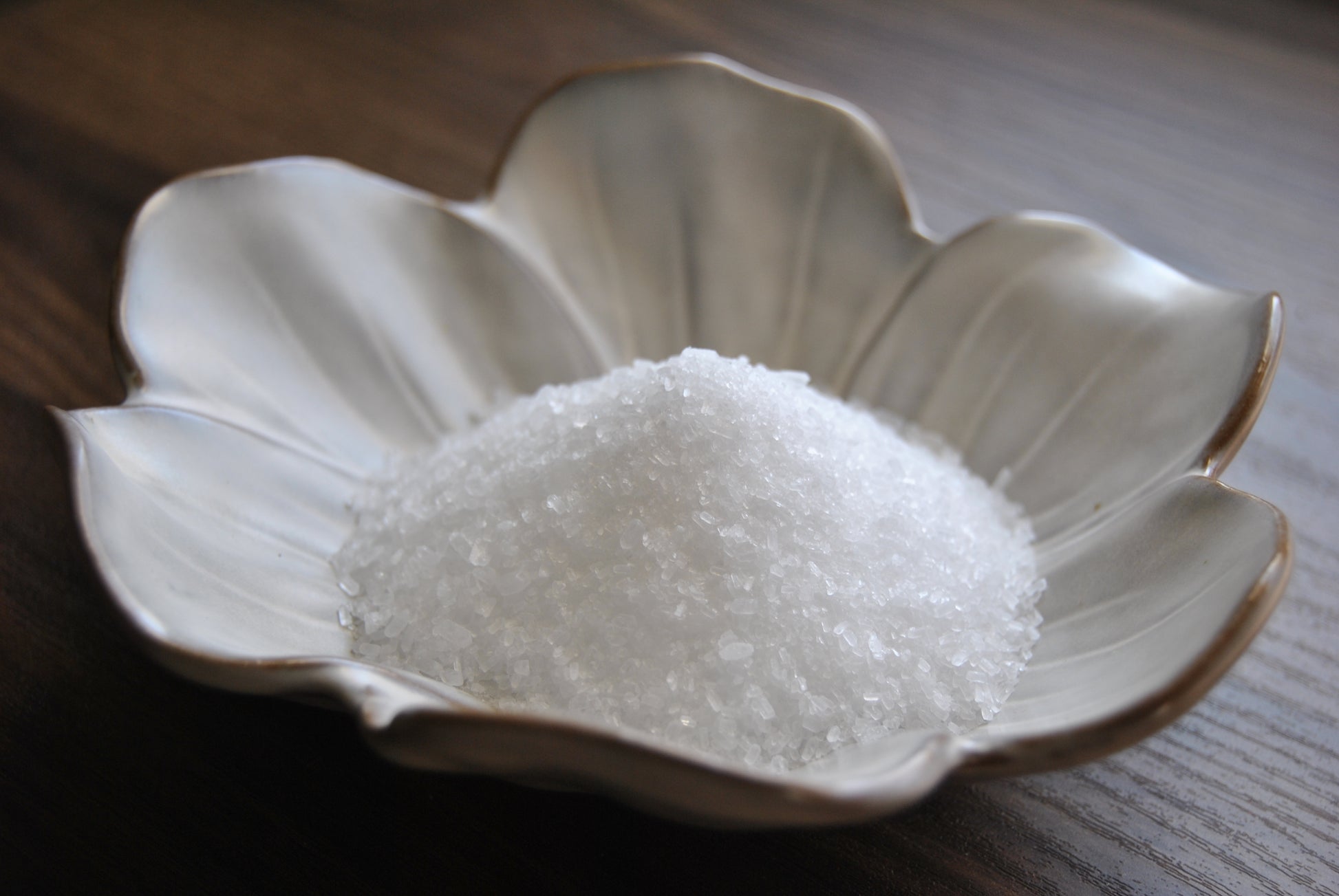 A close up view of the crystalline and clear epsom salt rests in a white, flower shaped dish on a wooden surface.