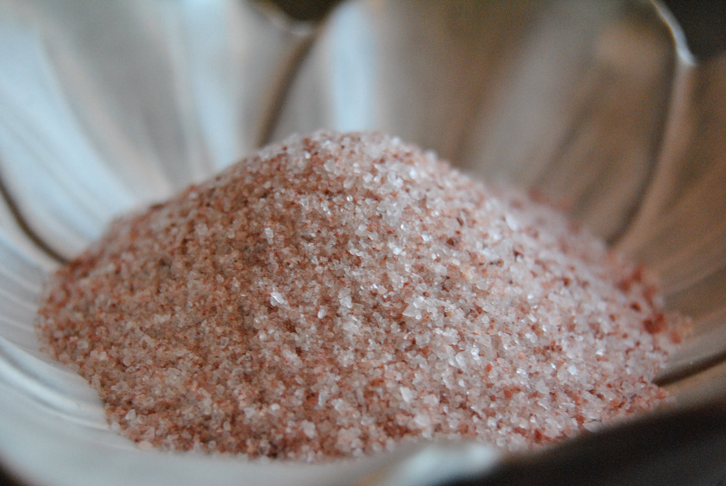 A pile of medium fine granules of pink himalayan salt rests in a white, flower shaped dish on a dark surface. The salt is light pink, with speckles of darker pink strewn throughout.