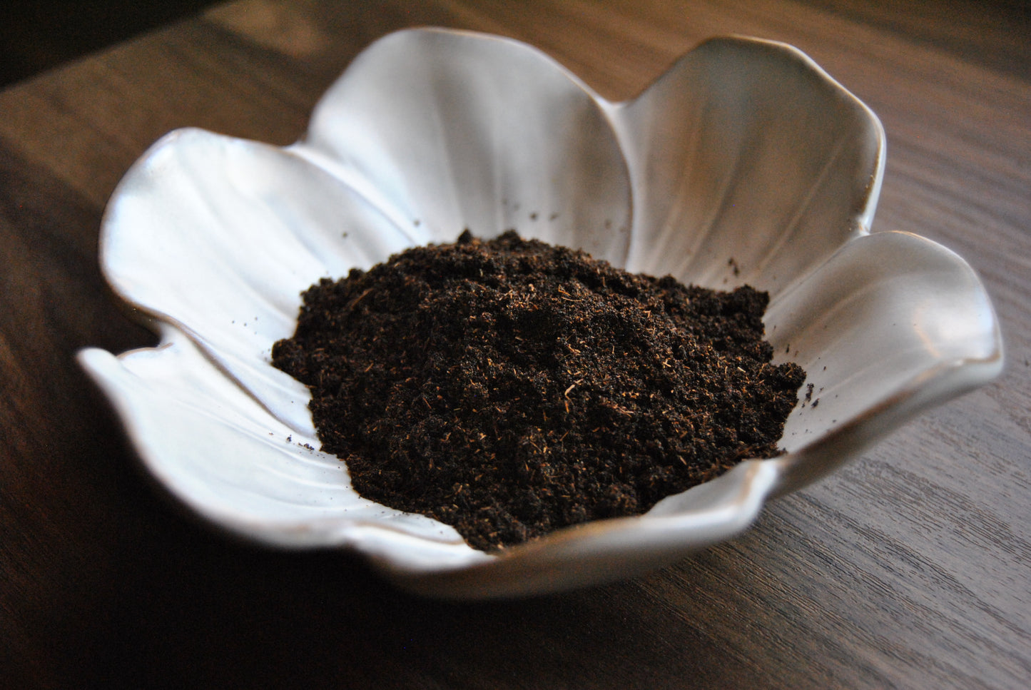 A pile vanilla powder rests in a white, flower shaped dish on a wooden surface. The powder is dark brown in color, with lighter speckles in it.