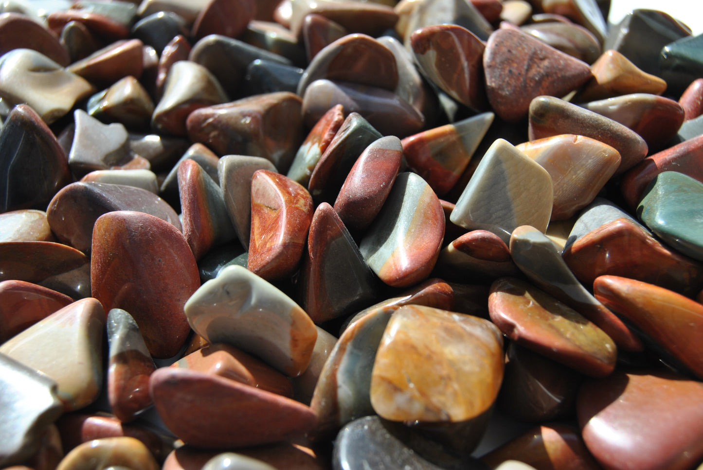 A close up view of an assortment of tumbled desert jasper stones. The stones vary in hues of red, orange, blue and brown.