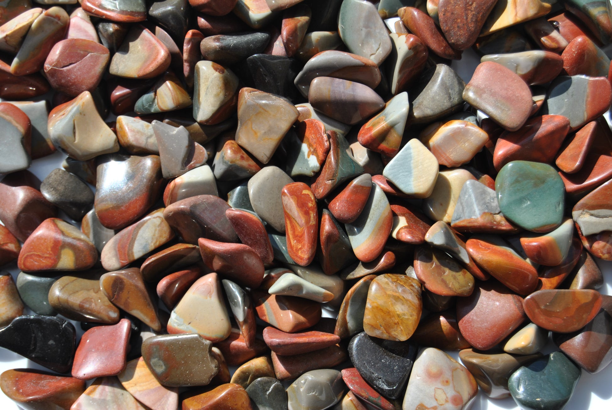 A top view of an assortment of tumbled desert jasper stones. The stones vary in hues of red, orange, blue and brown.