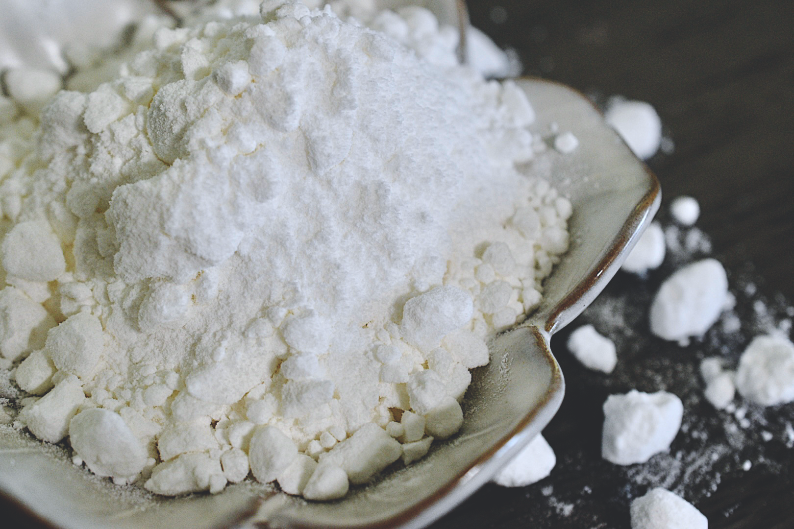 The white and silky coconut milk powder rests in a white, flower shaped dish on a wooden surface. Small clumps of powder are in the background of the photo.