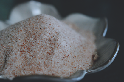 A pile of fine granules of pink himalayan salt rests in a white, flower shaped dish on a dark surface. The salt is light pink, with speckles of darker pink strewn throughout.