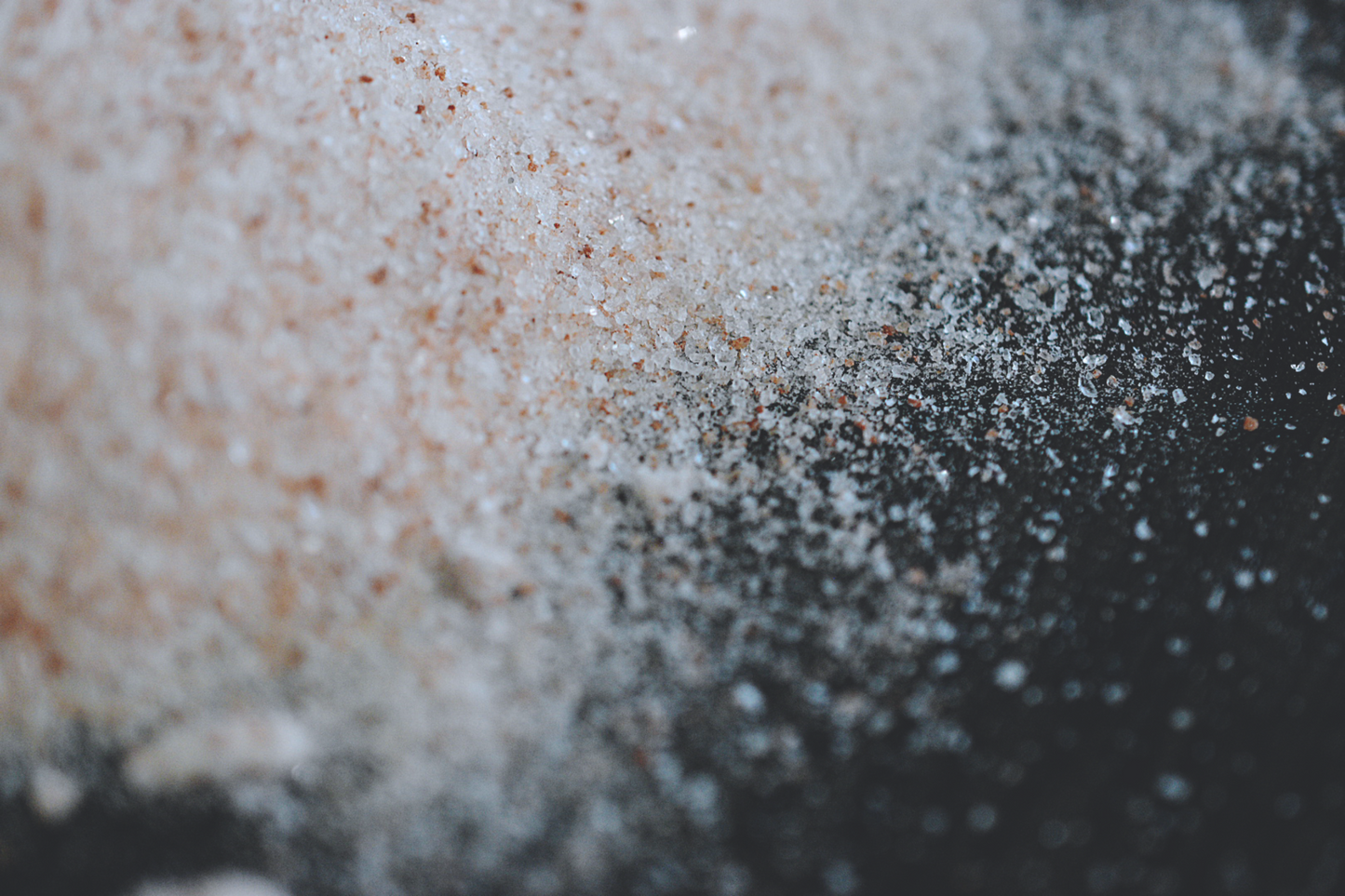 A close up view of the fine granules of pink himalayan salt scattered accross a dark surface. The pink salt is a light pink, with speckles of dark pink strewn throughout.