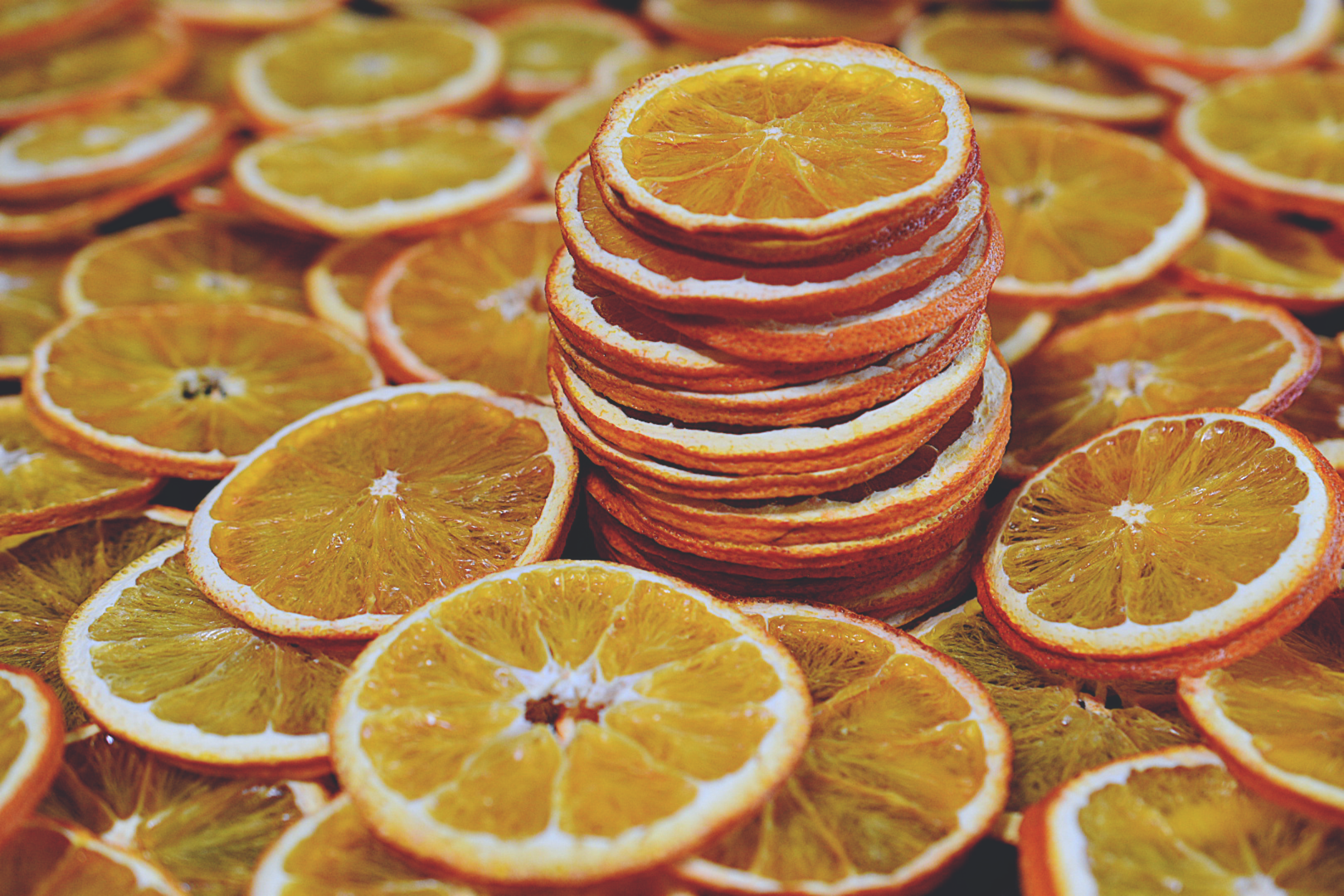 Many dried orange slices rests on a wooden surface. There is a stack of orange slices in the middle. The orange slices are deep orange in color.