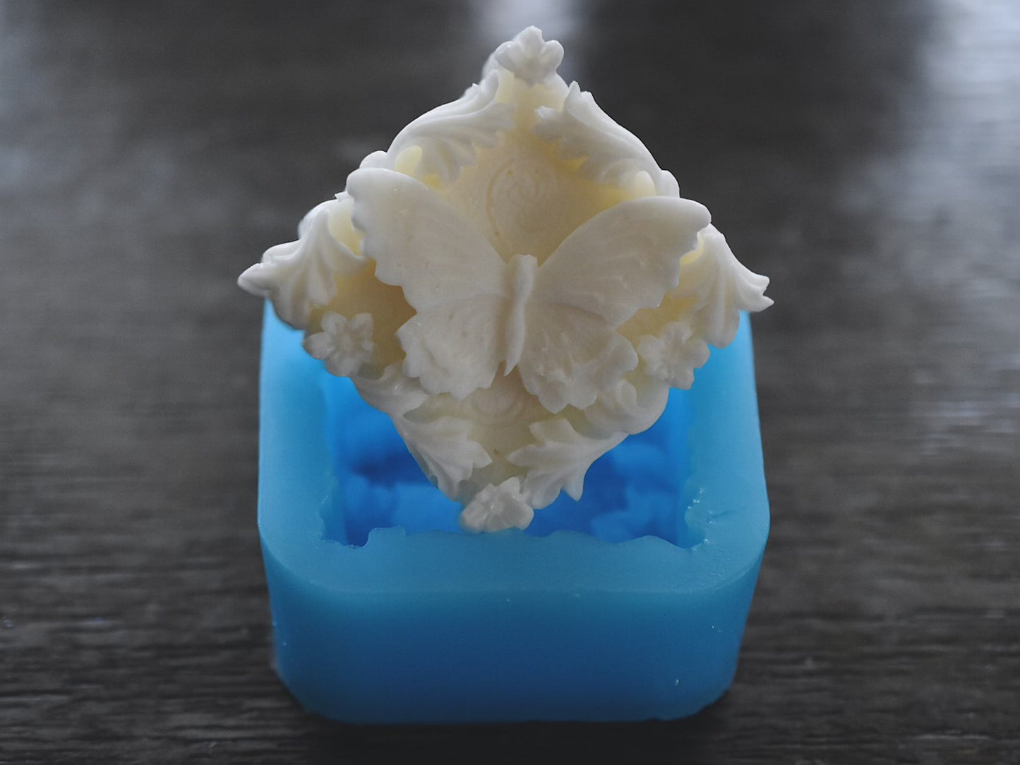 A square bar of soap with a flower and butterfly design sits on top of the butterfly mold on a wooden surface. The soap bar is white. The mold is blue.