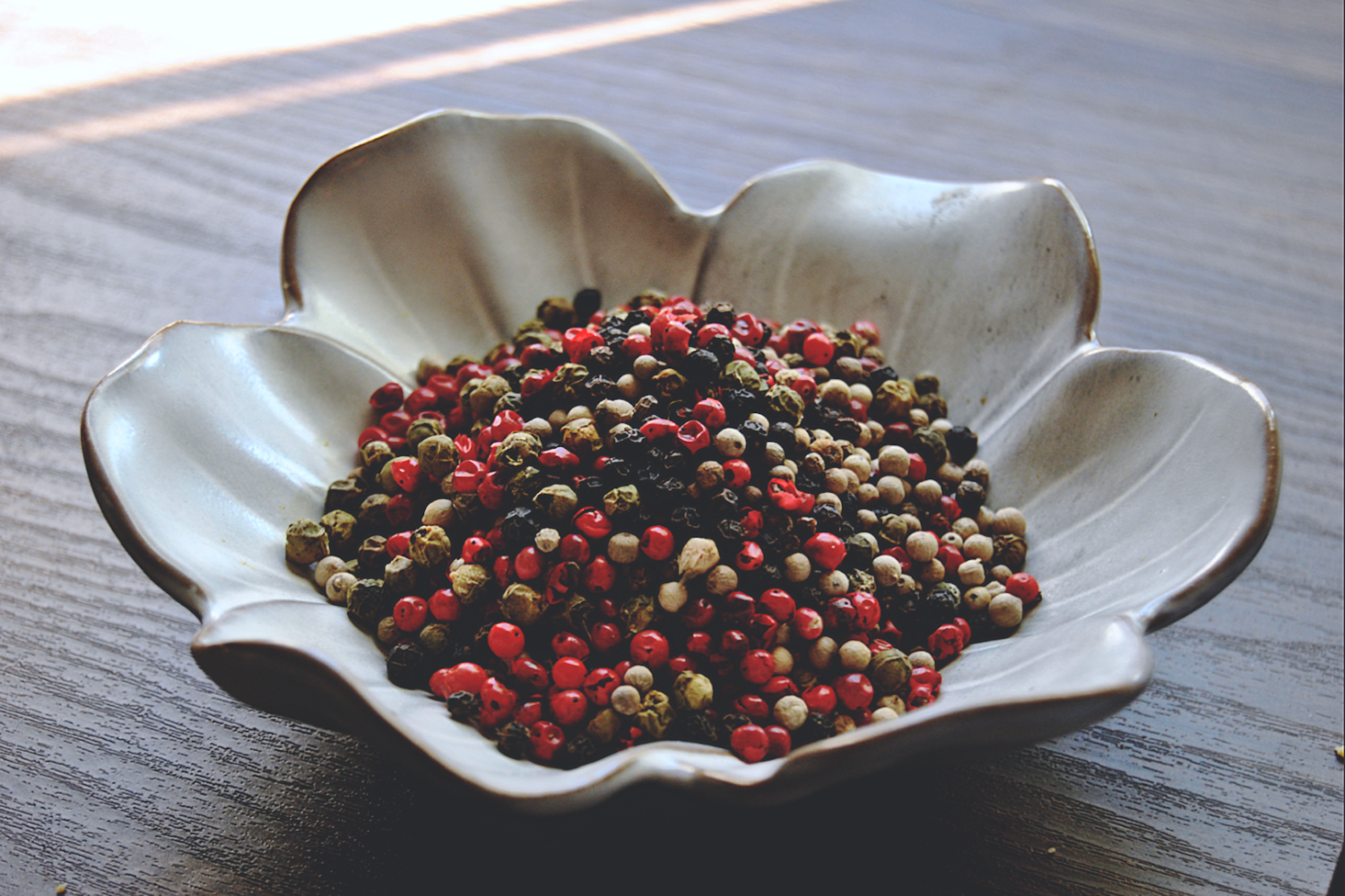 An assorted blend of pink, white, green and black peppercorns rests in a white, flower shaped dish on a wooden surface.
