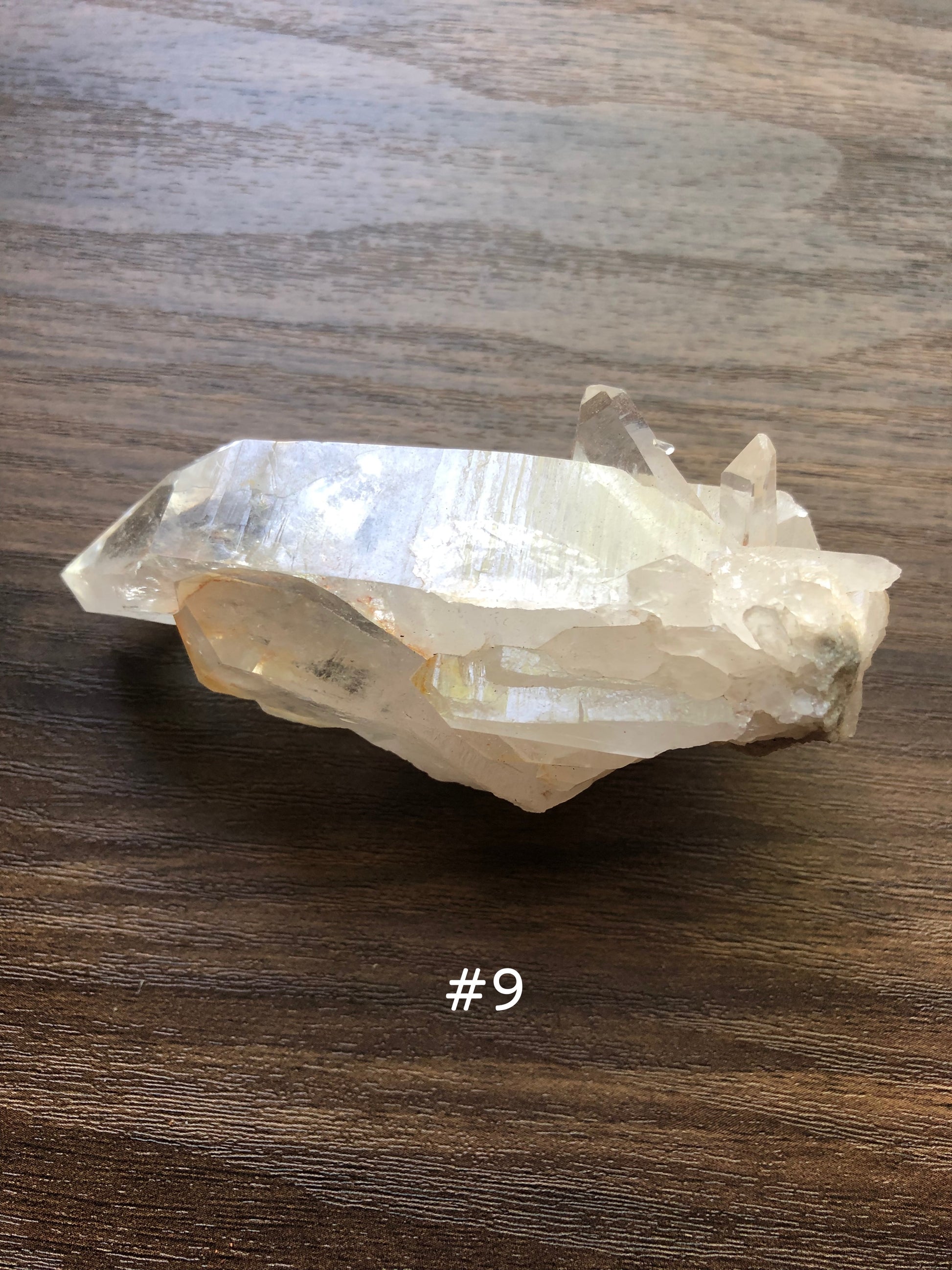 A large, rough cut quartz crystal rests on a wooden surface. It is jagged in shape with smaller clusters on it. The crystal is relatively clear with some slight discoloration. The number 9 is shown on the picture.