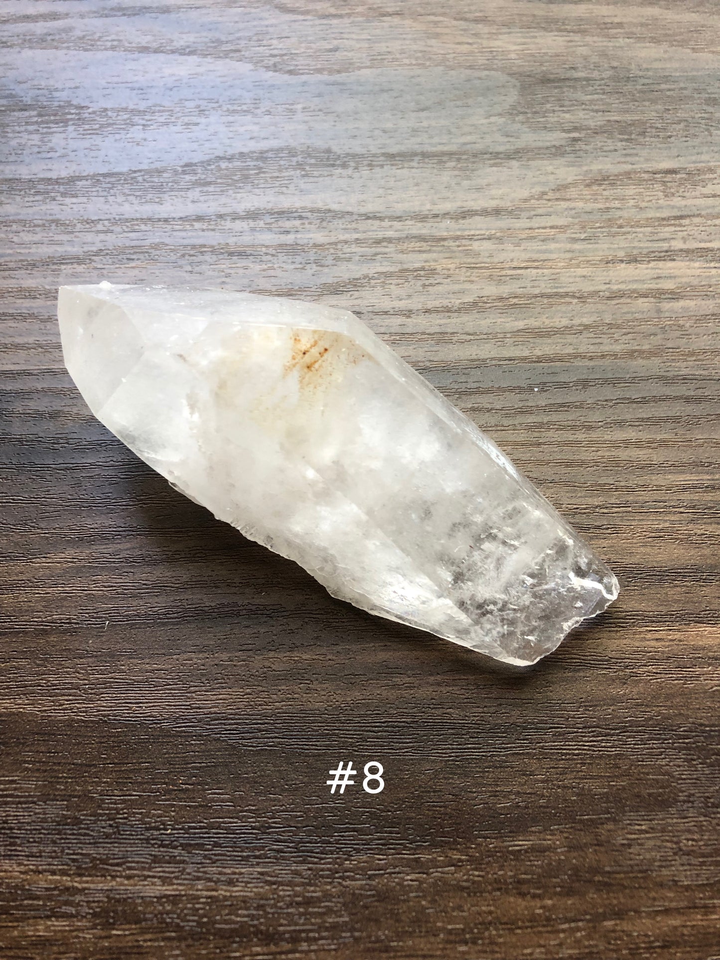 A large, rough cut quartz crystal rests on a wooden surface. It comes to a fine point, and is relatively flat.  The crystal is relatively clear, with a small dark spot on it. The number 8 is shown on the picture.