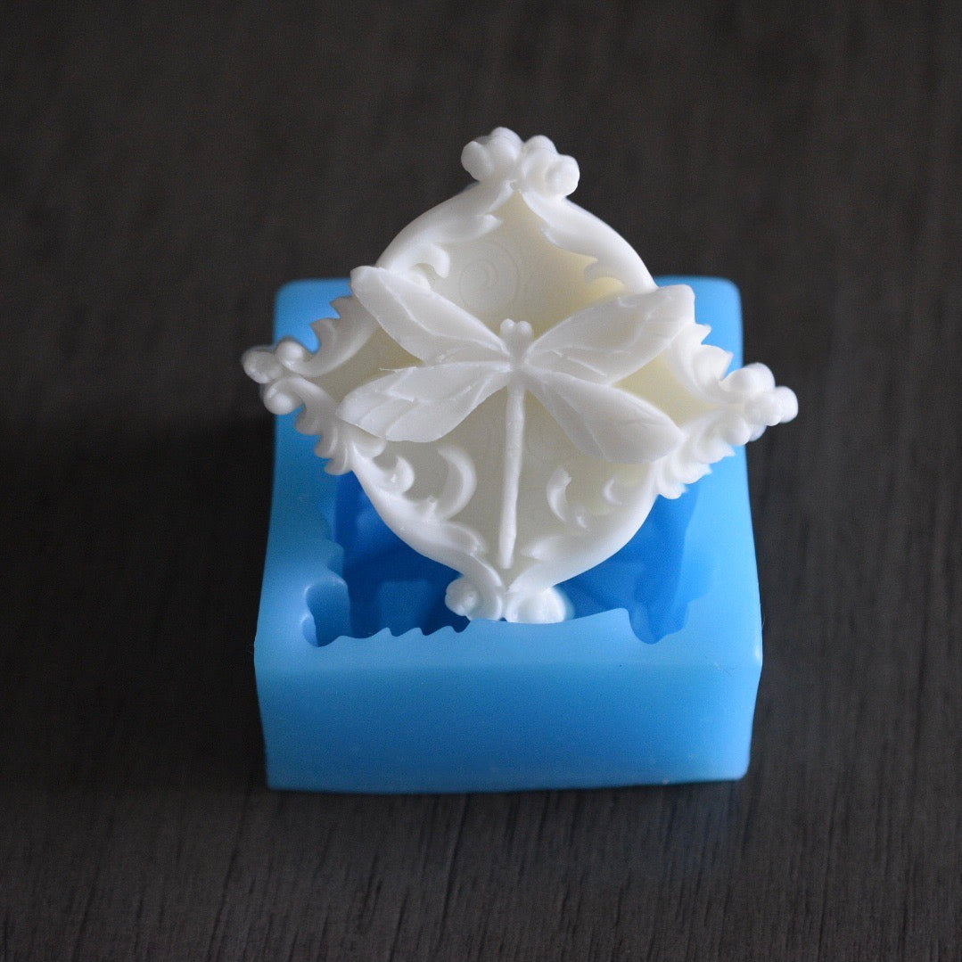 A square soap bar with a dragonfly design rests in a square mold on a wooden surface. The soap bar is white. The mold is blue.