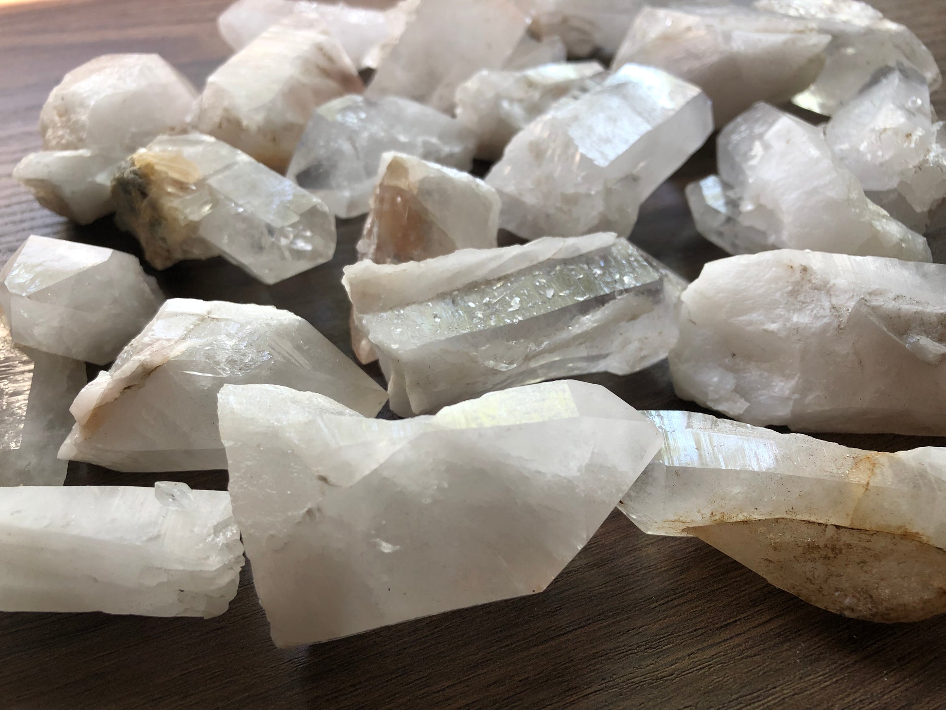 A close up view of an assortment of medium sized rough cut quartz crystals on a wooden surface. They are various shapes. They are all relatively clear, with some having slight discoloration.