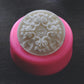 A round bar of soap with an elegant, filigree design rests in a round soap mold on a wooden surface. The mold is pink.