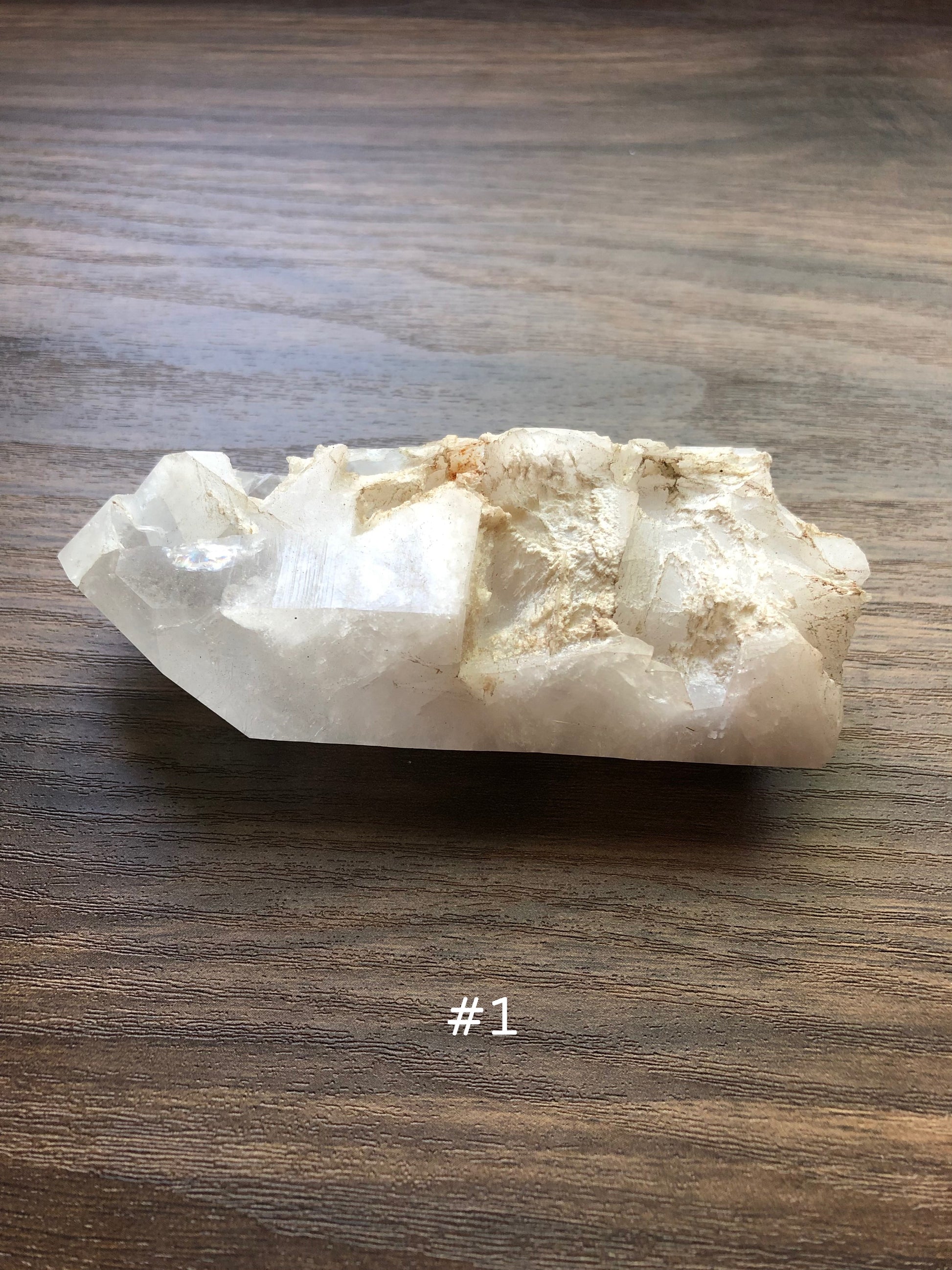 A large, rough cut quartz crystal rests on a wooden surface. It is jagged in shape. The crystal is relatively clear, with part of it being discolored. The number 1 is shown on the picture.