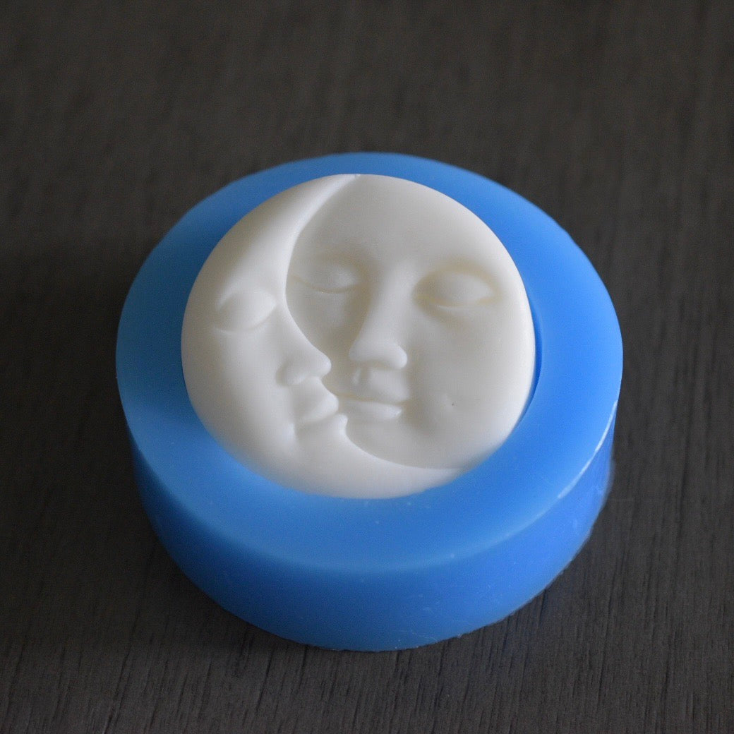 A round sun and moon soap bar that depicts the sun and moon with stoic faces sits in a sun and moon mold on a wooden surface. The sun and moon are face to face. The soap is colored white. The mold is blue.