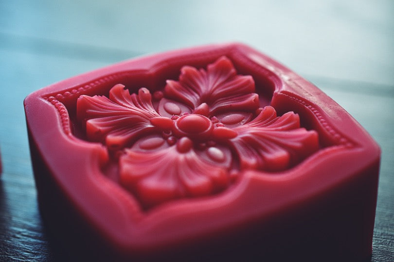 A close up of a square soap bar with a floral design rests on a wooden surface. The soap bar is colored a deep red.