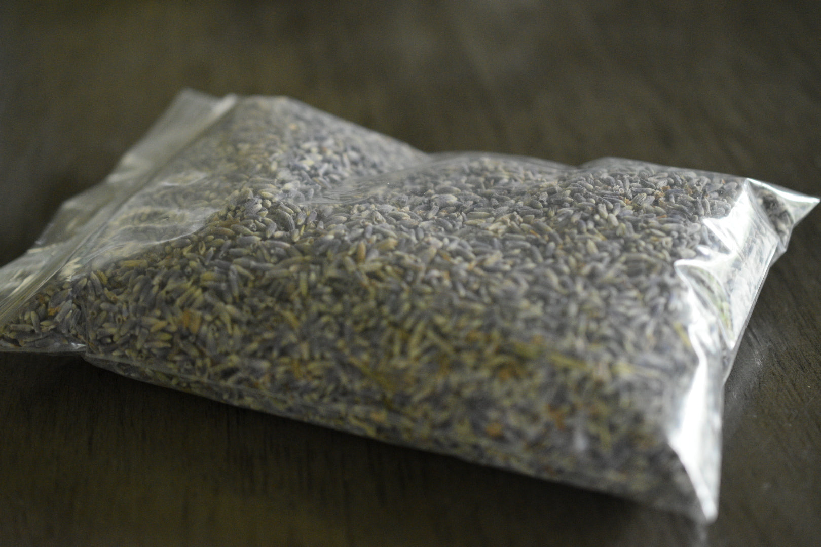 A plastic bag filled with lavender petals rests on a wooden surface to show the packaged product. The lavender is a soft purple with hints of blue.