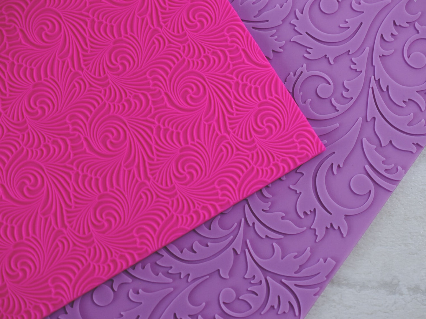 A close up view a textured pink mat with swirls and a textured purple mat with a floral filigree design are resting on a wooden surface. The pink mat is resting on the purple mat slightly.