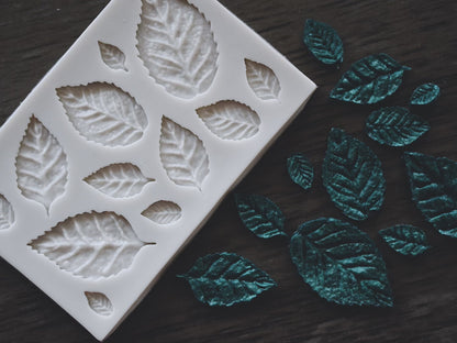An assortment of beech leaves made from soap next to a small beech leaf mold on a wooden surface. The beech leaves are colored green. The mold is grey.
