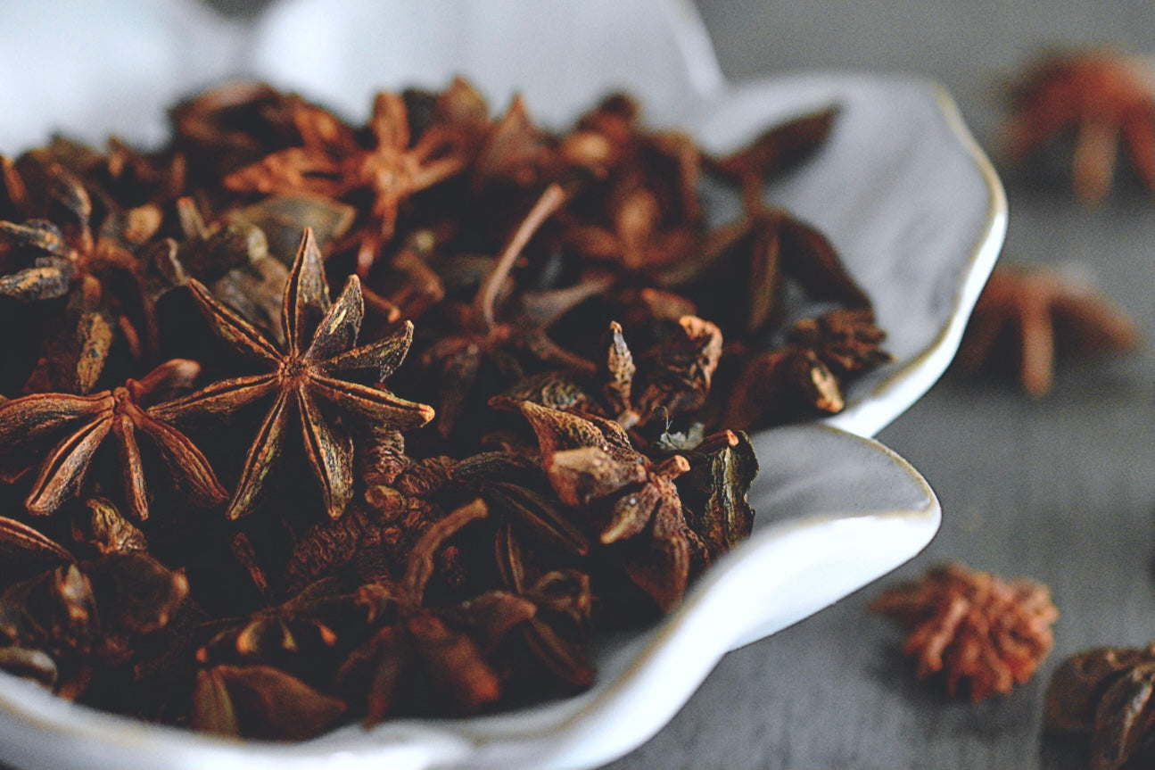 A close up view of an assortment of dried star anise flowers rests in a white, flower shaped bowl on a wooden surface. The dried flowers are dark brown in color. Some are chipped and have petals that are missing. Some flowers are scattered about in the background.