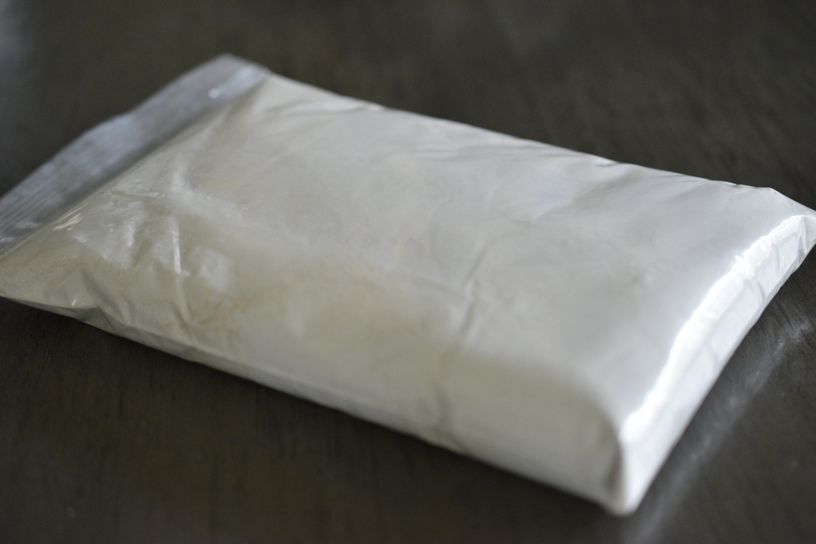 A plastic bag filled with kaolin clay rests on a wooden surface to show the packaged product. The clay is grey.