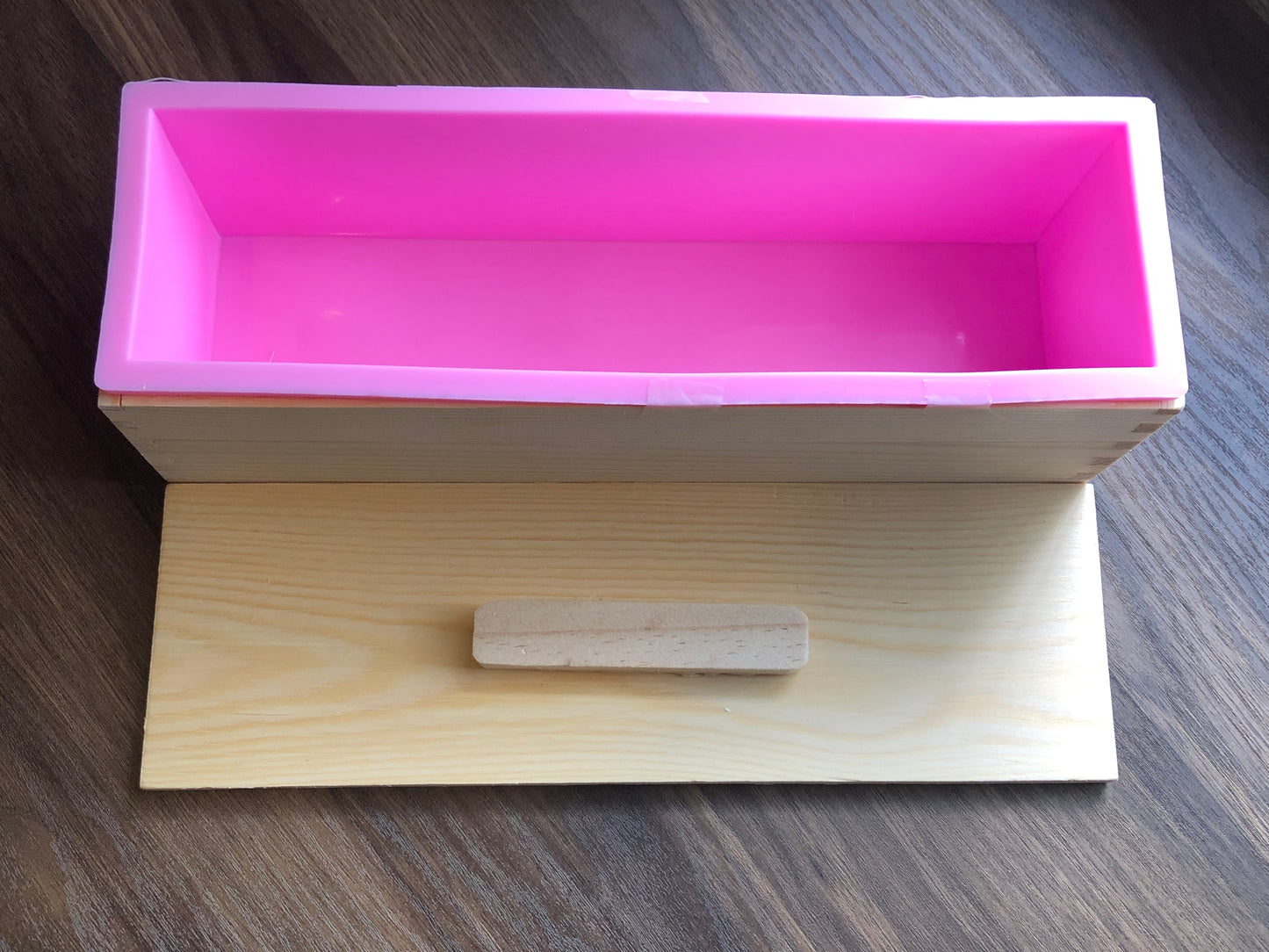A loaf shaped wood box that houses a pink silicone mold inside rests on a wooden surface. The lid is off to the side, showing the inside of the loaf mold. The lid has a handle. The wood of the mold is light in color.