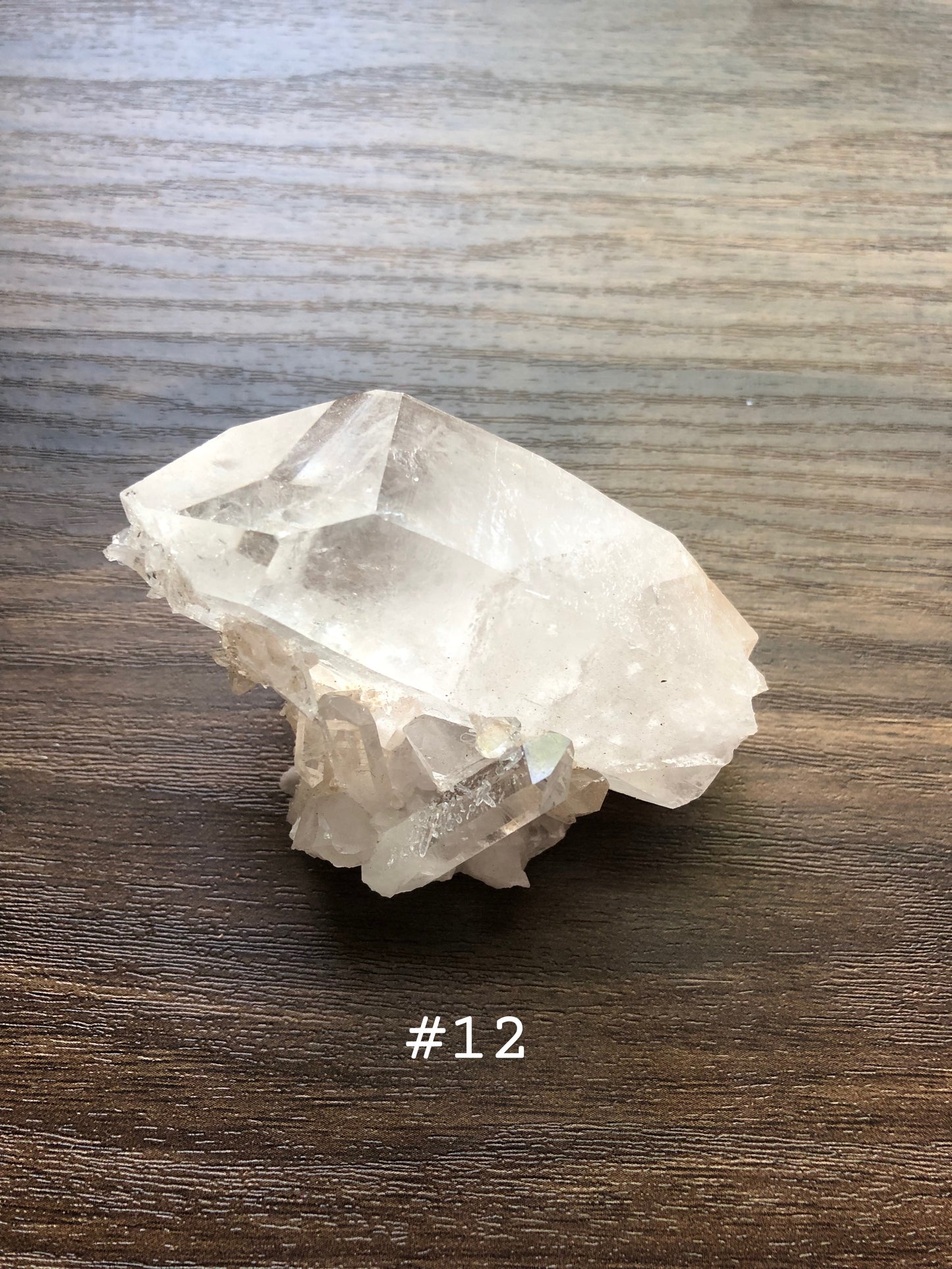 A large, rough cut quartz crystal rests on a wooden surface. It is jagged, with small clusters on it. The crystal is relatively clear. The number 12 is shown on the picture.