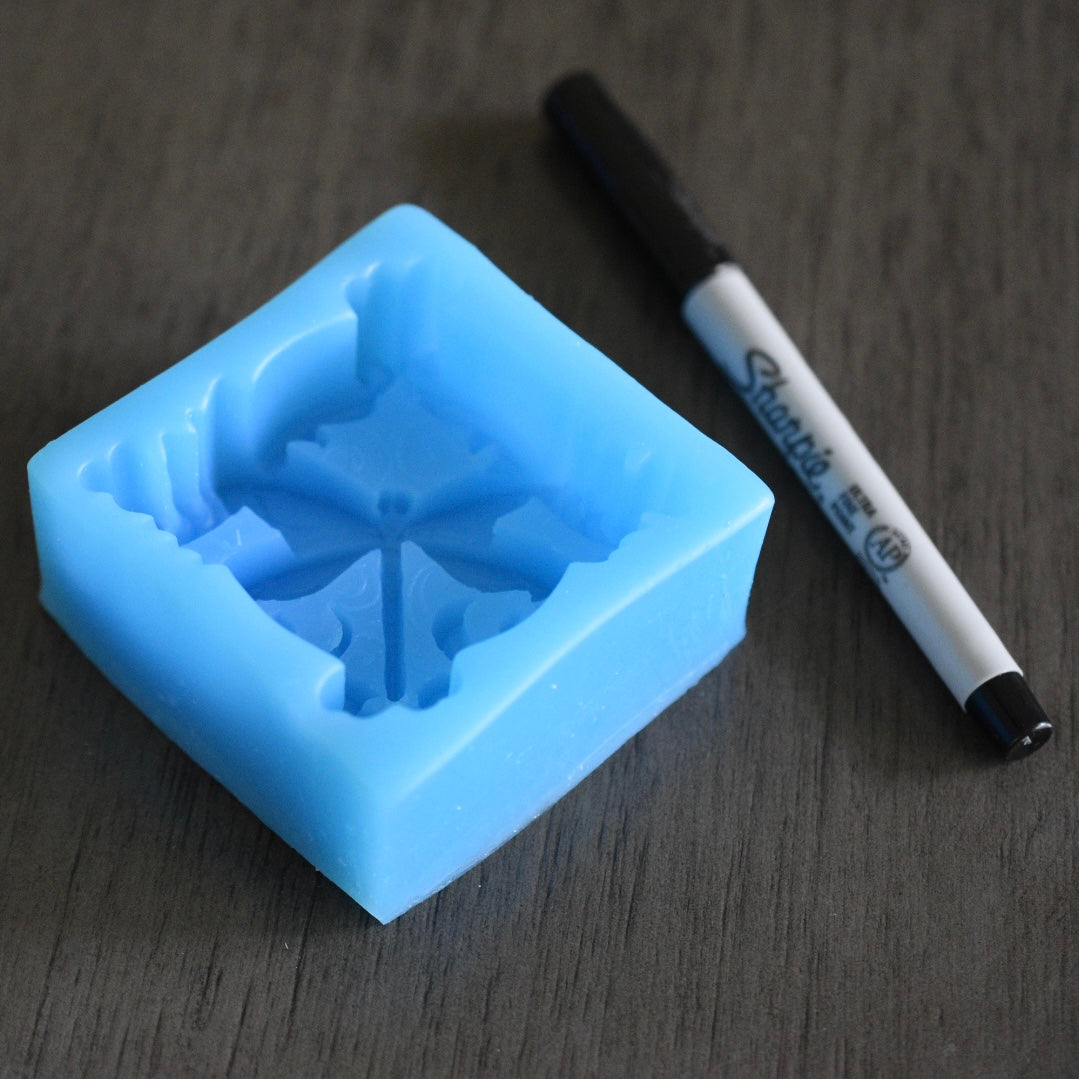 A square soap mold with a dragonfly design rests next to a sharpie pen on a wooden surface for size comparison. The sharpie pen is longer than the mold in length. The mold is blue.