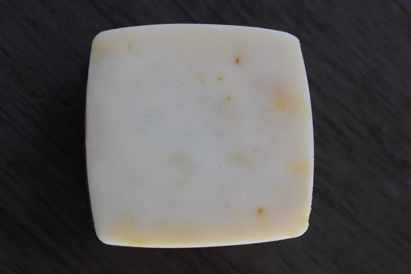 A top down view of a square bar of soap on a wooden surface. The bar is white, with speckles of orange and yellow color in it from the orange powder.