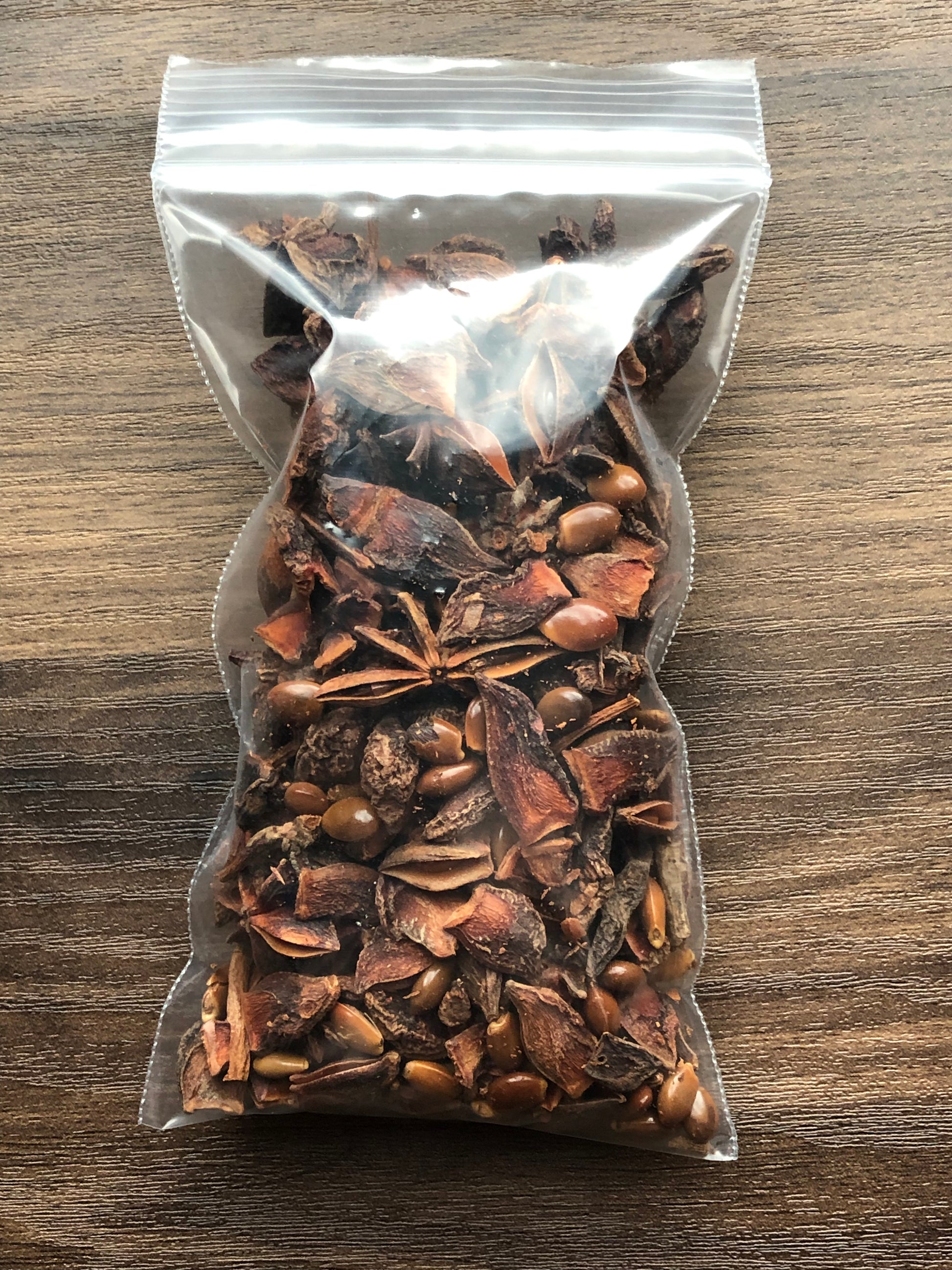 A small plastic bag filled with star anise pieces to show the packaged product rests on a wooden surface. Most pieces are missing petals or are chipped. The star anise pieces are dark brown in color.
