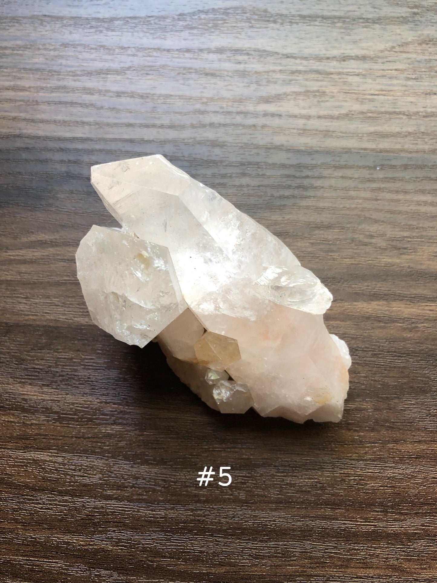 A large, rough cut quartz crystal rests on a wooden surface. It is jagged in shape with smaller crystals coming out of it. The crystal is relatively clear. The number 5 is shown on the picture.
