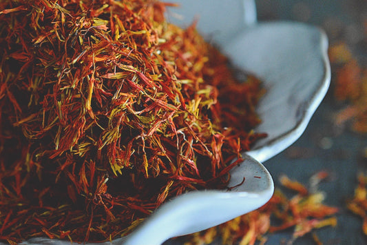 A close up view of an assortment of safflower petals rests in a white, flower shaped dish on a wooden surface. The safflower petals are a vibrant red with hints of orange. Saffllower petals are scattered about in the background.