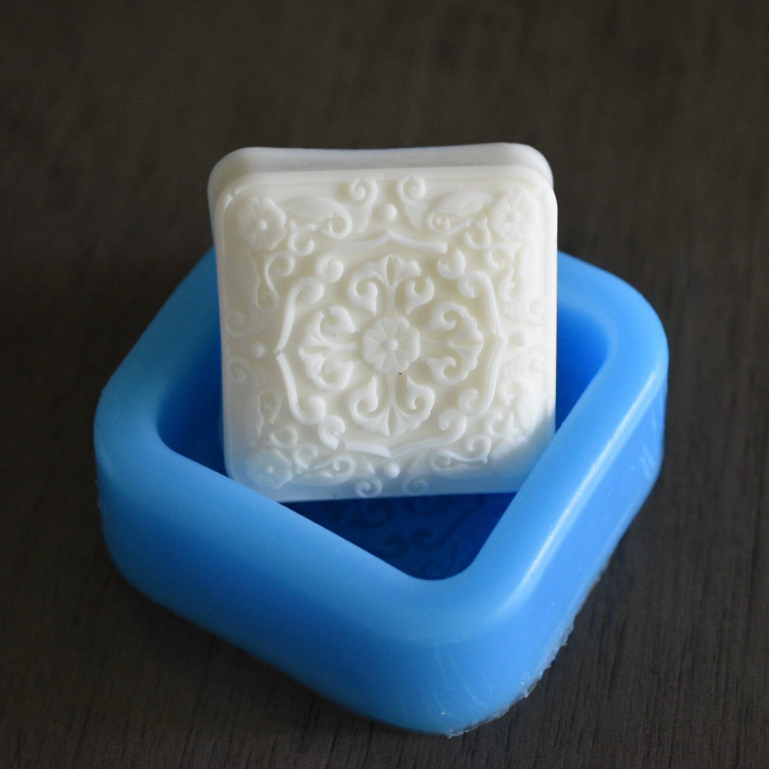 A square soap bar with a filigree design rests in a square mold on a wooden surface. The soap bar is white. The mold is blue.