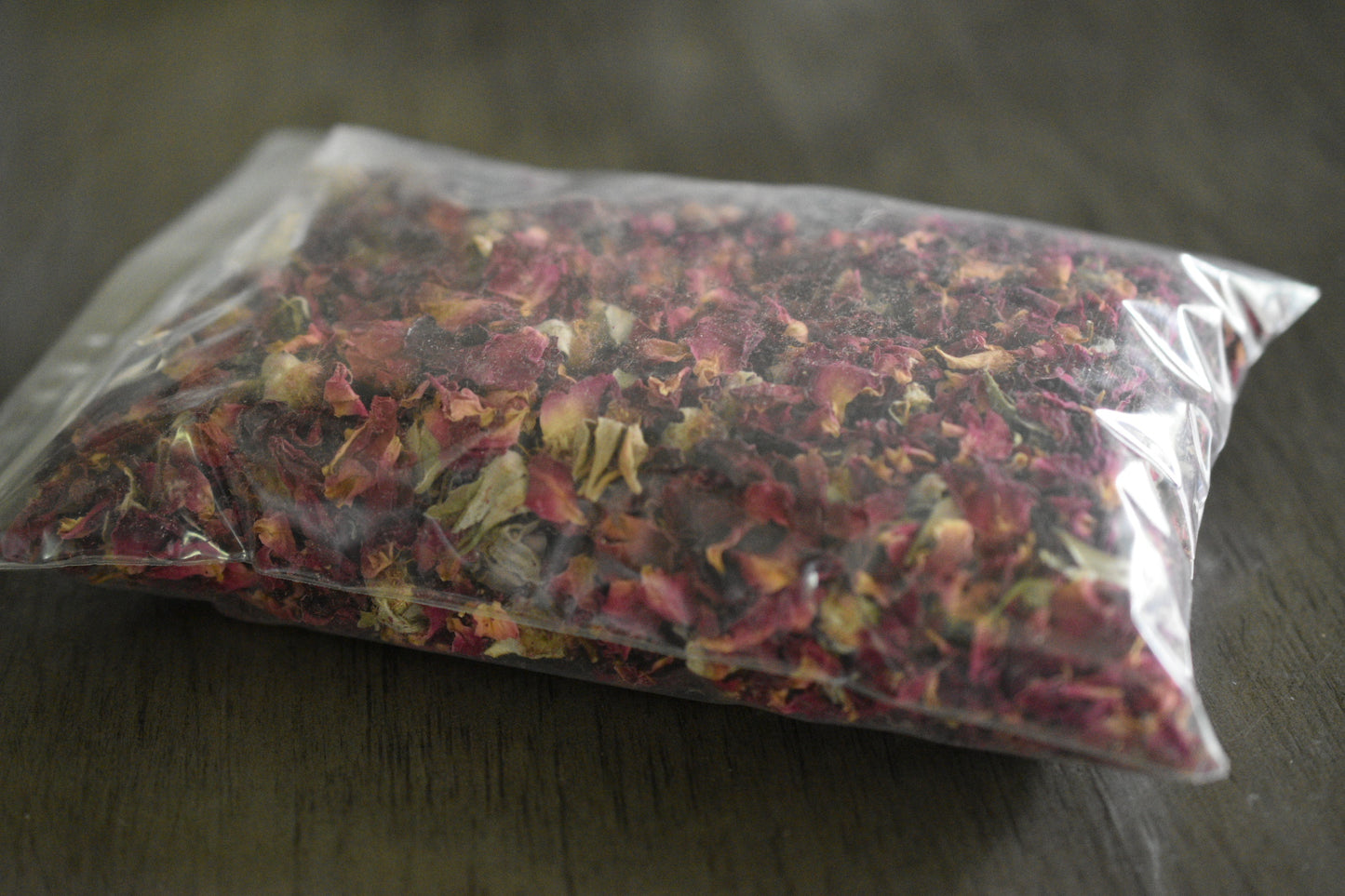 A plastic bag filled with rose petals and buds rests on a wooden surface to show the packaged product. The rose is a deep pink and red color.