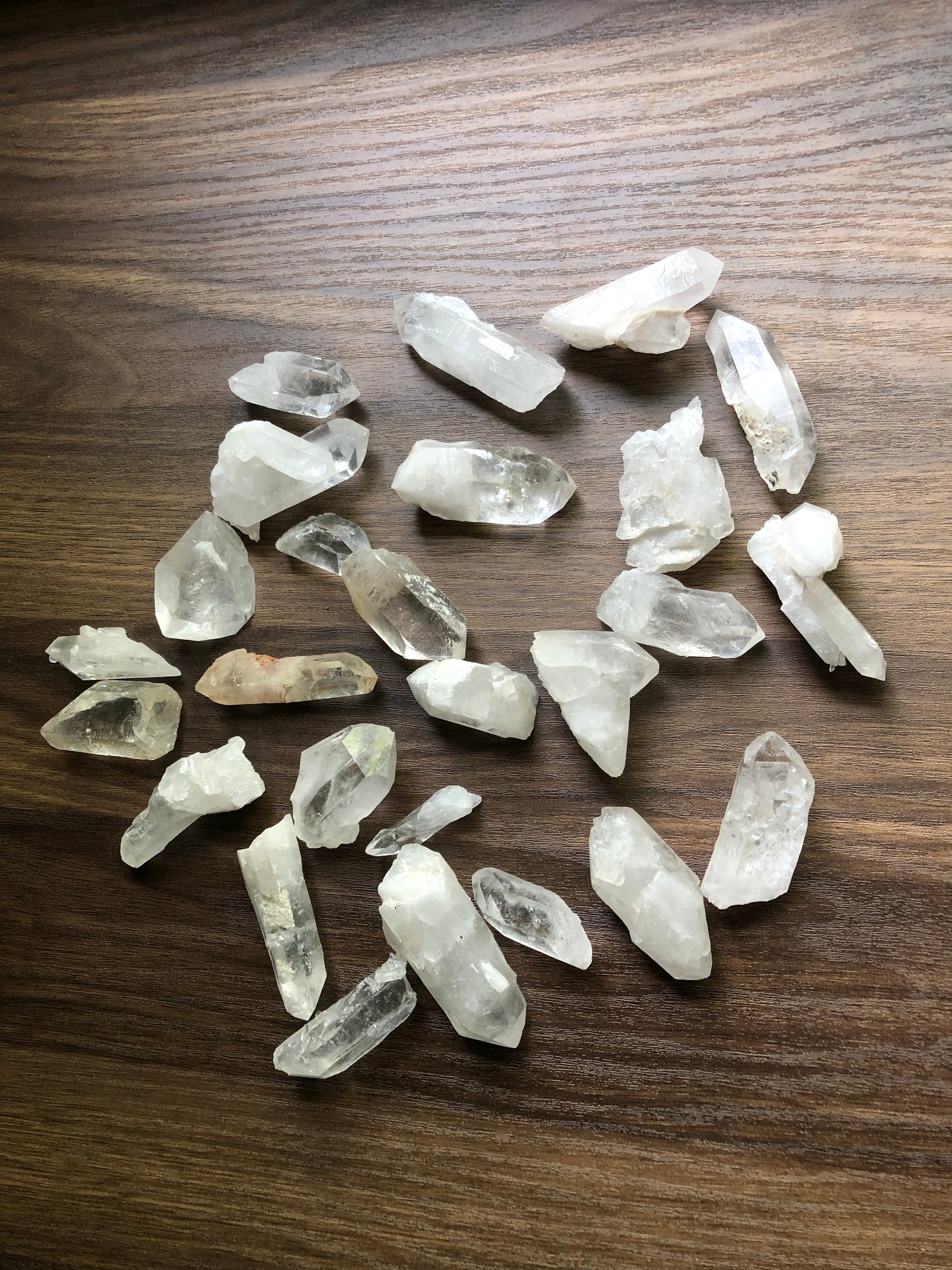 A top down view of an assortment of small sized rough cut quartz crystals on a wooden surface. They are various shapes. They are all relatively clear, with some having slight discoloration.