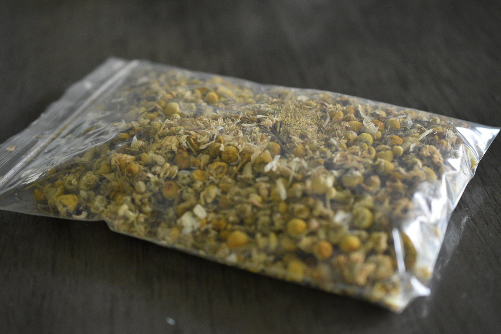 A plastic bag filled with light yellow chamomile buds rests on a wooden surface to show the packaged product.