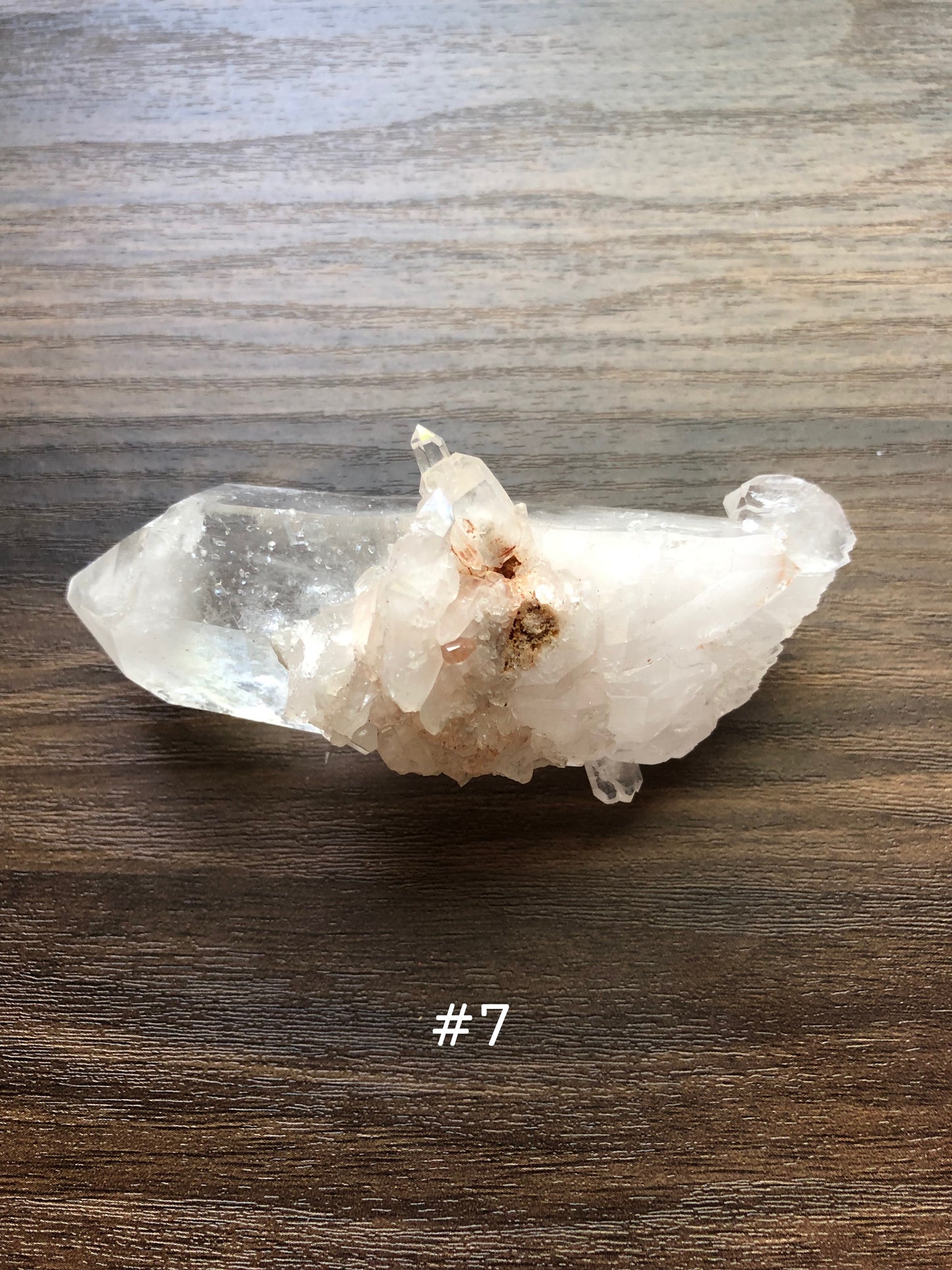A large, rough cut quartz crystal rests on a wooden surface. It is jagged, with small clusters on it.  The crystal is relatively clear, with the clusters being slightly discolored. The number 7 is shown on the picture.