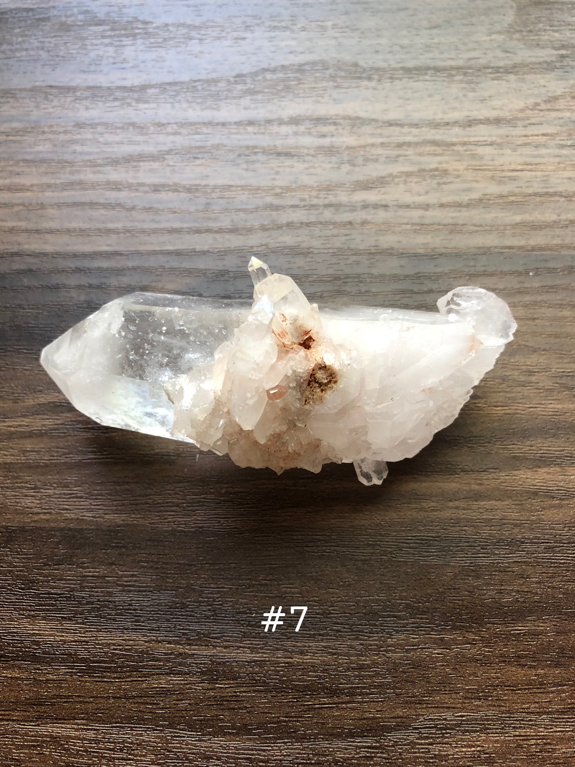 A large, rough cut quartz crystal rests on a wooden surface. It is jagged, with small clusters on it.  The crystal is relatively clear, with the clusters being slightly discolored. The number 7 is shown on the picture.