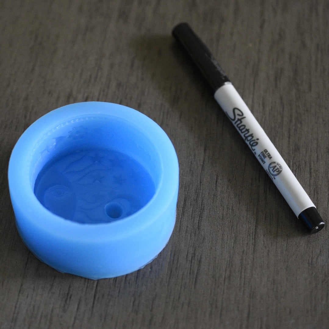 A round soap mold rests next to a sharpie pen on a wooden surface for size comparison. The mold is smaller in length than the pen. The mold is blue.