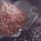 A pile of coarse granules of pink himalayan salt rests in a white, flower shaped dish on a dark surface. The salt is light pink, with speckles of darker pink strewn throughout.