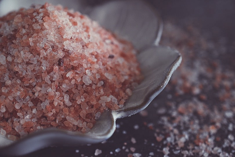 A pile of coarse granules of pink himalayan salt rests in a white, flower shaped dish on a dark surface. The salt is light pink, with speckles of darker pink strewn throughout.