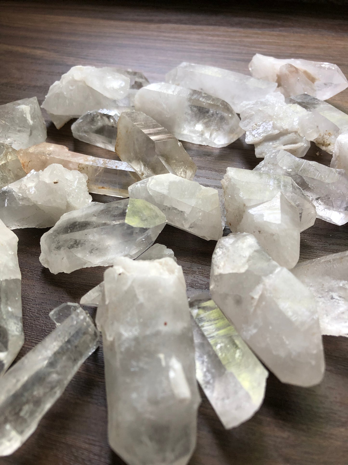 A close up view of an assortment of small sized rough cut quartz crystals on a wooden surface. They are various shapes. They are all relatively clear, with some having slight discoloration.