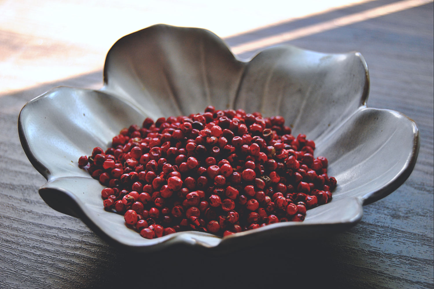 An assortment of pink peppercorns rests in a white, flower shaped dish on a wooden surface.
