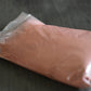 A plastic bag filled with rose clay rests on a wooden surface to show the packaged product. The clay is light pink in color.