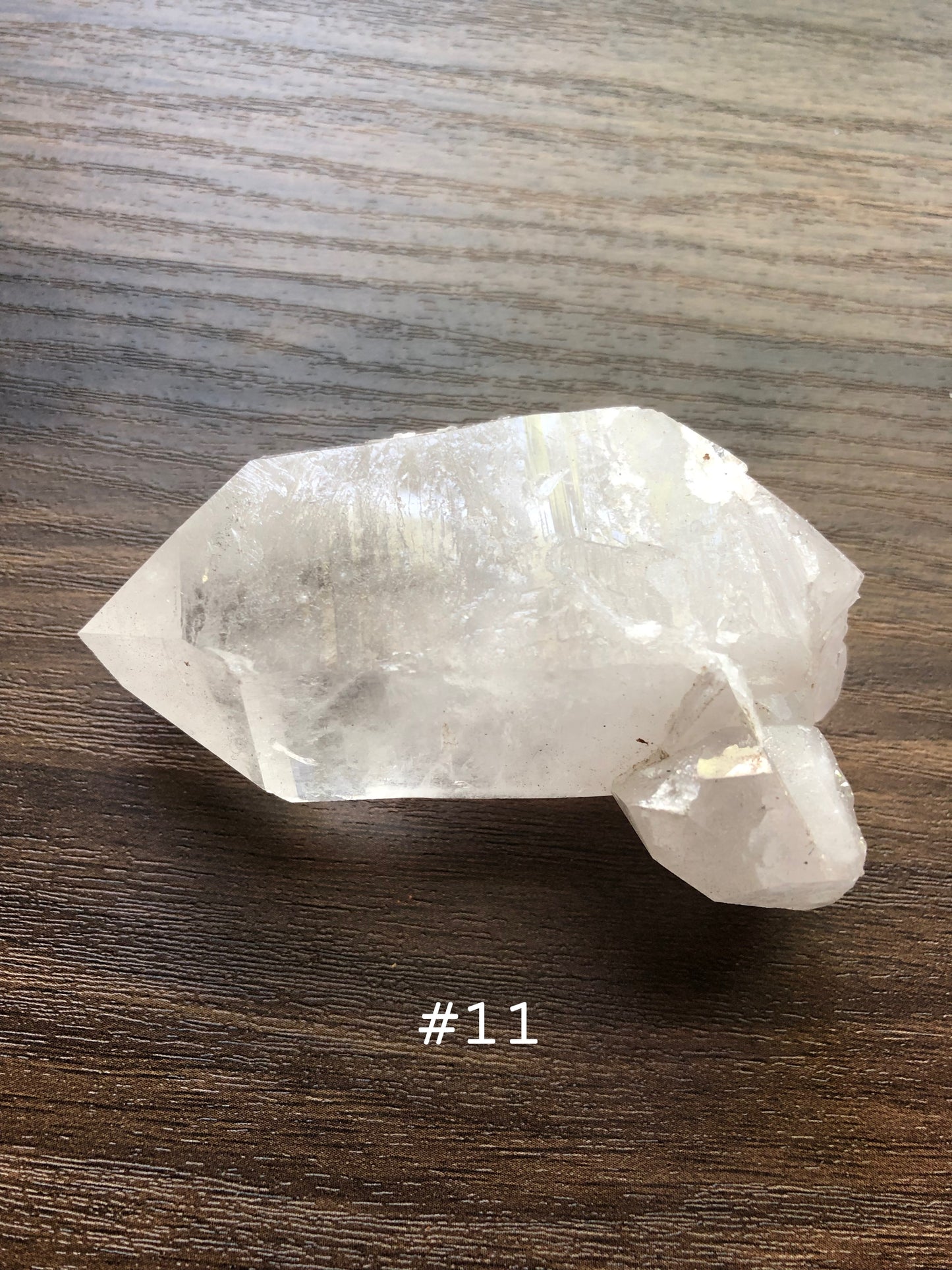 A large, rough cut quartz crystal rests on a wooden surface. It comes to a small point with a small cluster on it. The crystal is relatively clear. The number 11 is shown on the picture.