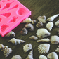 An assortment of seashells made from soap rests next to a square soap mold on a wooden surface. The seashells are colored a cream and brown color. The mold is pink.