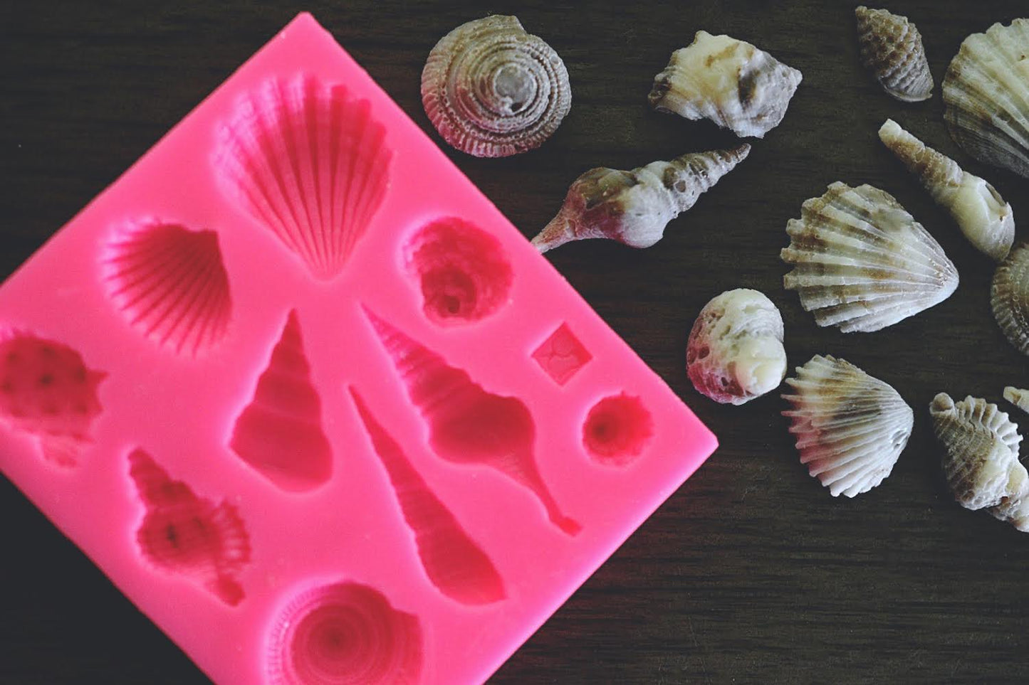 A close up view of an assortment of seashells made from soap rests next to a square soap mold on a wooden surface. The seashells are colored a cream and brown color. The mold is pink.