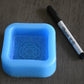 A square soap mold with a filigree design rests next to a sharpie pen on a wooden surface for size comparison. The sharpie pen is longer than the mold in length. The mold is blue.