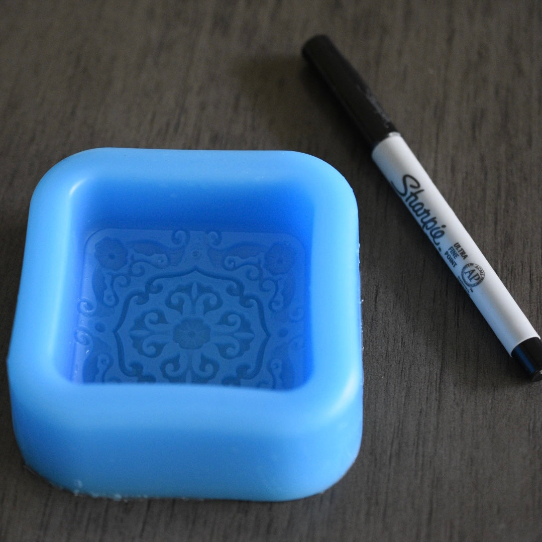 A square soap mold with a filigree design rests next to a sharpie pen on a wooden surface for size comparison. The sharpie pen is longer than the mold in length. The mold is blue.