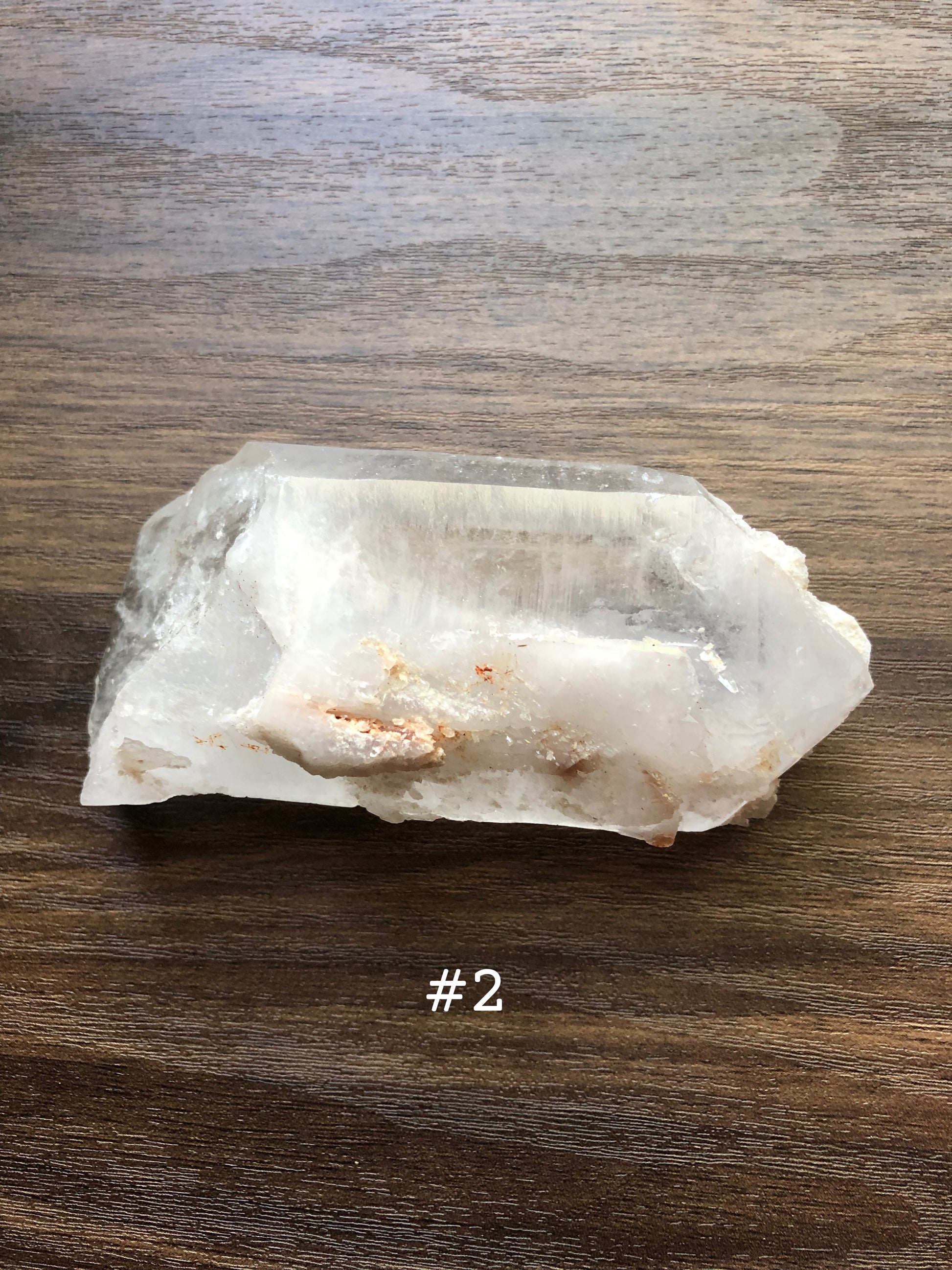 A large, rough cut quartz crystal rests on a wooden surface. It is jagged in shape. The crystal is relatively clear, with a portion of it being slightly discolored. The number 2 is shown on the picture.